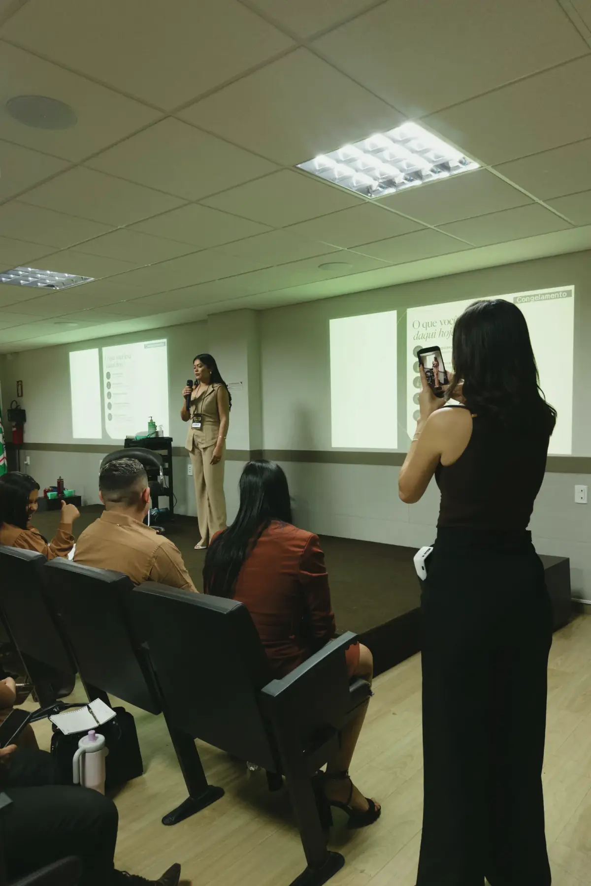 Pâmella em pé, filmando uma palestra com o celular, com a plateia em primeiro plano.