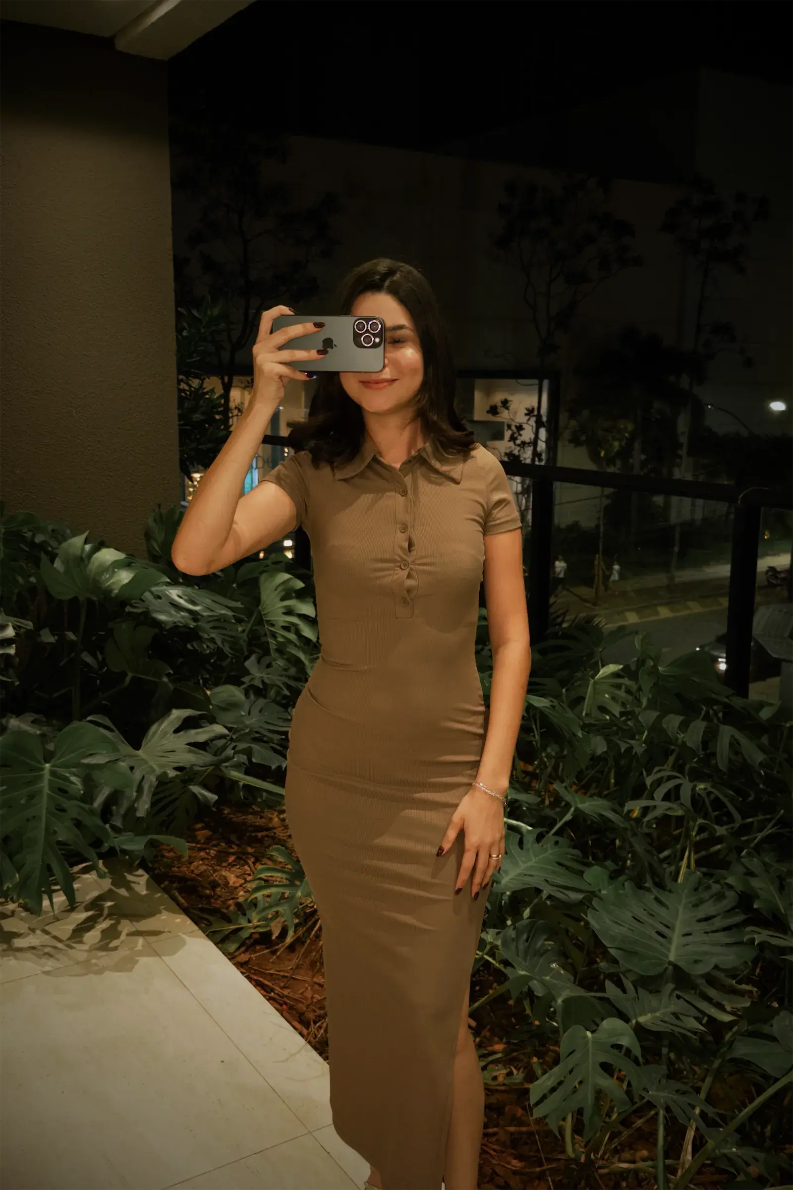 Pâmella de vestido caramelo num jardim à noite, segurando o celular na frente do rosto como quem grava.